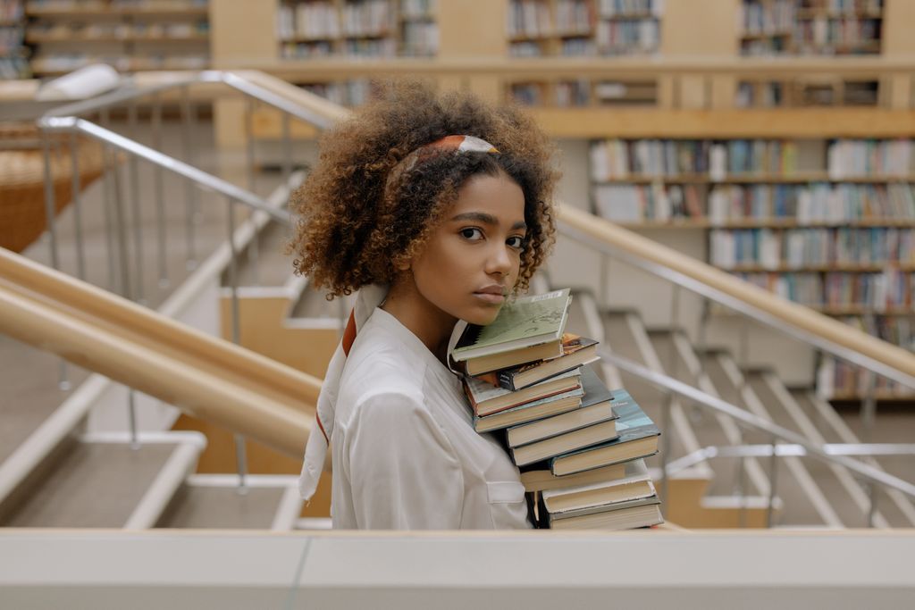 A young woman with curly hair holding a stack of books in a library.